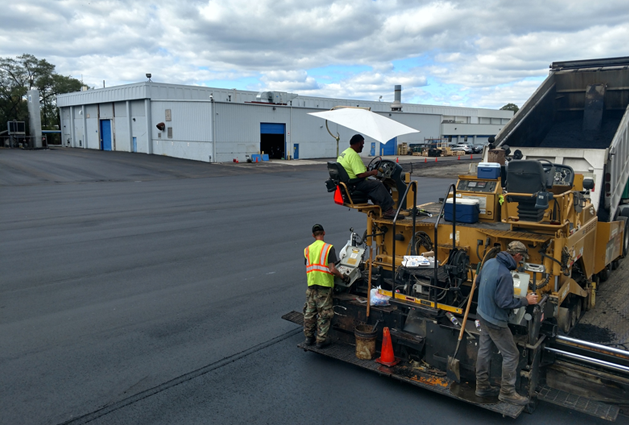 paving contractor crew on a yellow asphalt laying vehicle paving a parking lot in front of a white warehouse